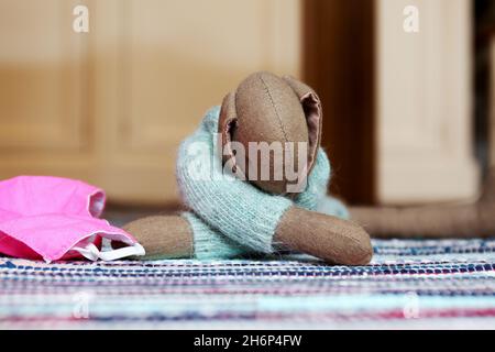 Kuscheltier Süßer Hase mit langen Ohren und hellblauem Wollpulli, liegend auf einem bunten Teppich. Daneben eine rosa farbene Maske (Mundschutz). Foto Stock