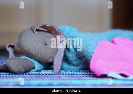 Kuscheltier Süßer Hase mit langen Ohren und hellblauem Wollpulli, liegend auf einem bunten Teppich. Daneben eine rosa farbene Maske (Mundschutz). Foto Stock