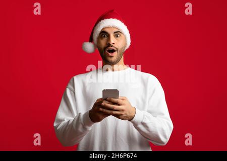 Offerta incredibile. Ritratto di uomo arabo sorpreso in smartphone Santa Hat Holding Foto Stock