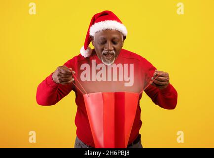 Uomo americano africano anziano in Santa Hat guardando all'interno della borsa shopping con luce magica, godendo il suo regalo di Natale Foto Stock