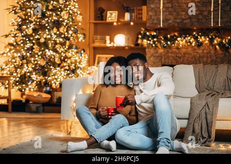 Allegra giovane coppia nera che festeggia il Natale a casa, seduto sul pavimento con un caffè, trascorrere il tempo insieme a Natale Foto Stock