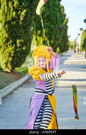 Felice clown femminile con volto dipinto in costume divertente e parrucca gialla, sorridente e camminando sul sentiero nel parco con poi colorato nelle mani durante la sfilata Foto Stock