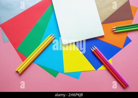 Concetti di scuola e hobby. Fogli di carta colorati, matite colorate e libro di schizzo aperto su sfondo di carta rosa. Spazio libero per testo o foto Foto Stock