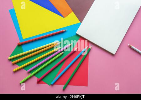 Concetti di scuola e hobby. Fogli di carta colorati, matite colorate e libro di schizzo aperto su sfondo di carta rosa. Spazio di copia Foto Stock