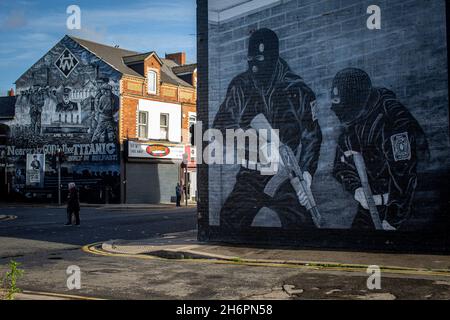 Persone che camminano davanti a un murale paramilitare lealista con uomini mascherati che tengono mitragliatrici sulla Newtownards Road a Belfast, Irlanda del Nord. Foto Stock