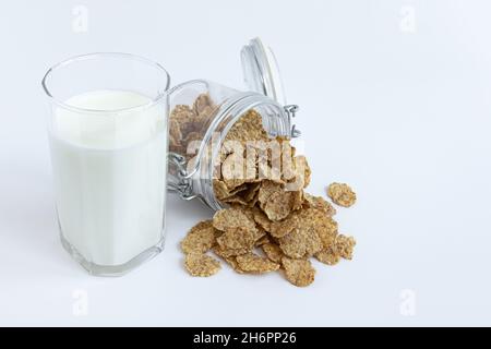 vaso di vetro con coperchio aperto si trova su sfondo bianco, fiocchi di grano intero per una colazione sana caduta da esso, vetro trasparente con latte fresco biologico a. Foto Stock