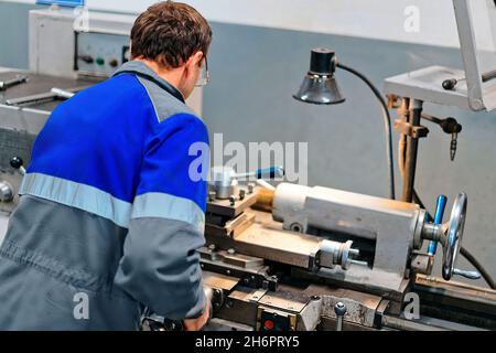 Il torcente adulto di 50-55 anni lavora in officina dietro il tornio. Produzione industriale di parti metalliche. Lavoratore caucasico in tute. Vista posteriore. Autentico processo di lavoro scena in produzione. Lavoratore reale. Scena non in scena. Foto Stock