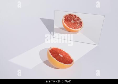 Composizione alla moda e minimal fatta di pompelmo rosso e il suo riflesso specchio su elegante sfondo bianco. Creativo concetto di frutta estiva. Foto Stock
