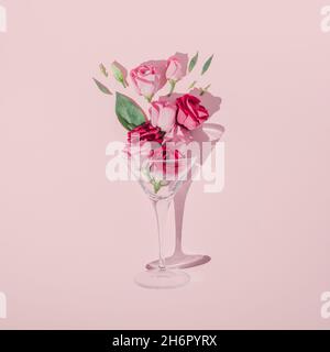 Composizione romantica di San Valentino realizzata con bicchiere da cocktail, foglie verdi e rose rosa su sfondo pastello. Disposizione piatta minima. Foto Stock