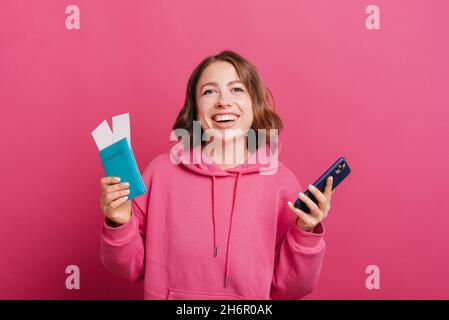 Giovane e allegra sorridente donna con passaporto con biglietti e smartphone, pronta per il viaggio. Foto Stock