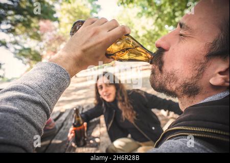 Giovane coppia che beve birra dalla bottiglia su panca di legno con tavolo nel parco forestale all'aperto. Espresso bere e veloce ottenere ubriaco stile di vita concetto. Reale Foto Stock