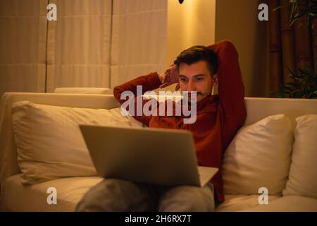 Un giovane uomo con una barba in una camicia rossa lavora con un computer portatile e si siede sul divano la sera in casa. Foto Stock