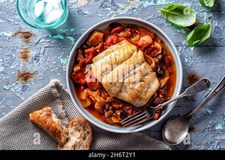 Un piatto di delizioso pesce fritto in padella in stile mediterraneo con pomodoro, finocchio, cipolla e olive kalamata su un piano da tavola rustico. Foto Stock