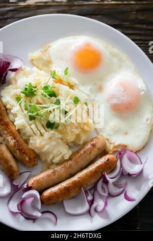 Salsicce di maiale con cipolle, uova, purè di patate e microgreens. Colazione abbondante. Sfondo in legno scuro, piastra in ceramica bianca. Vista dall'alto. Foto Stock