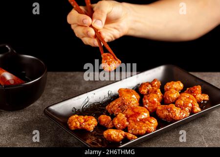 Una persona sta mangiando il pollo d'arancia del mandarino delizioso, un piatto americano cinese usando i bastoncini di legno. La salsa viene servita in una ciotola di porcellana. bac. Scuro Foto Stock