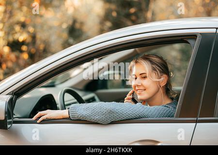 Foto di giovane bella donna bionda che parla sul finestrino dell'automobile aperto dello smartphone mentre si ferma il veicolo, sorridendo. Buon viaggio gioioso, momento di relax Foto Stock