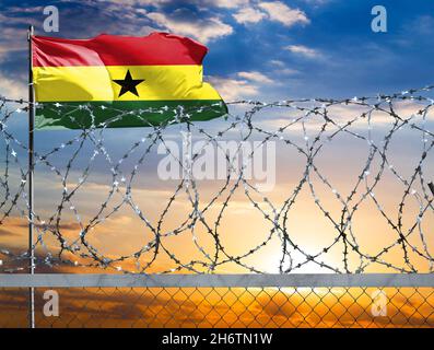 Una recinzione con filo spinato sullo sfondo di un cielo colorato e un flagpole con la bandiera del Ghana protegge il confine dello stato dalla migrazione illegale Foto Stock