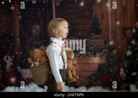 Carino moda bambino ragazzo, giocando nella neve con orsacchiotto di fronte a una cabina di legno di tronchi, decorazione di natale Foto Stock