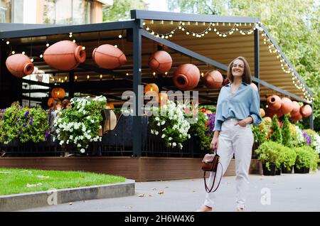 Ritratto urbano di giovane donna elegante in occhiali e abiti casual. Donna che cammina nella strada della città vicino alla fontana, rilassante, sorridente Foto Stock
