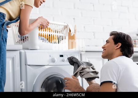 uomo sorridente che carica lavatrice vicino alla ragazza allegra afroamericana con cestino di lavanderia sporca Foto Stock