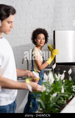 sorridente donna afroamericana che tiene la spugna vicino al frigorifero vicino uomo sfocato annaffiare le piante in cucina Foto Stock