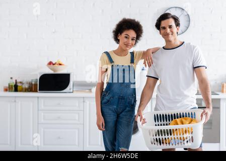 felice coppia interrazziale vicino cestino con lavanderia sporca Foto Stock