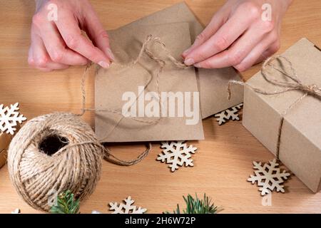 Le mani stanno avvolgendo i regali in uno stile rispettoso dell'ambiente. Regali fatti in casa, fai da te. Le mani legano un fiocco di spago su una busta con una lettera di Natale sul Th Foto Stock