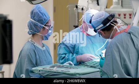 primo piano dei medici che operano su un paziente in una sala operatoria con uno studente o un infermiere che osservano, e un tavolo degli strumenti chirurgici accanto ad esso. Foto Stock