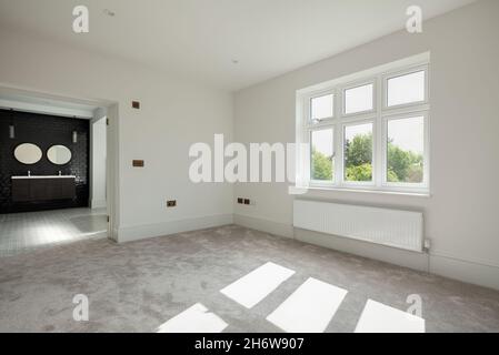 Wratting, Inghilterra - Agosto 19 2019: Camera da letto vuota e soleggiata con porte che si aprono su bagno privato piastrellato decorato semplicemente in bianco e moquette Foto Stock