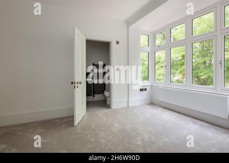 Wratting, Inghilterra - Agosto 19 2019: Camera da letto vuota con porta che si apre sul bagno ensuite semplicemente decorato in bianco con pavimento tappezzato Foto Stock