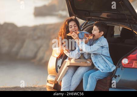 Una giovane madre e suo figlio adolescente in abiti denim seduti nel tronco aperto di una macchina e mangiare pizza Foto Stock