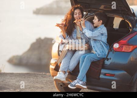 Una giovane madre e suo figlio adolescente in abiti denim seduti nel tronco aperto di una macchina e mangiare pizza Foto Stock