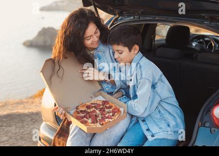 Una giovane madre e suo figlio adolescente in abiti denim seduti nel tronco aperto di una macchina e mangiare pizza Foto Stock