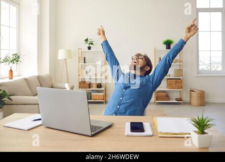 Un uomo d'affari felice di fronte al laptop che alza le mani e ride entusiasta del suo successo Foto Stock