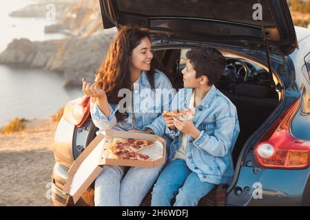 Una giovane madre e suo figlio adolescente in abiti denim seduti nel tronco aperto di una macchina e mangiare pizza Foto Stock