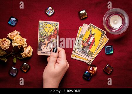 La mano del fortuneteller con manicure nero deposita le schede del tarot, il cristallo, la candela, le rose asciutte sul tablecloth rosso disposizione piatta in alto la vista della fortuna che dice, predire Foto Stock