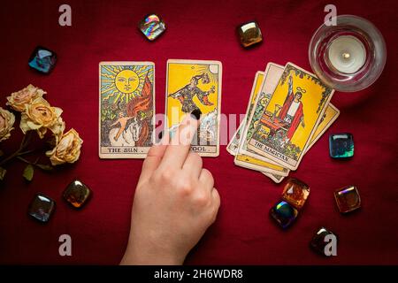 La mano del fortuneteller con manicure nero deposita le schede del tarot, il cristallo, la candela, le rose asciutte sul tablecloth rosso disposizione piatta in alto la vista della fortuna che dice, predire Foto Stock