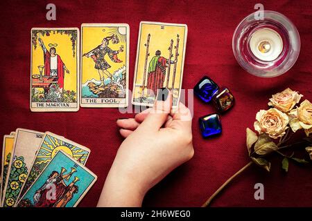 La mano del fortuneteller con manicure nero deposita le schede del tarot, il cristallo, la candela, le rose asciutte sul tablecloth rosso disposizione piatta in alto la vista della fortuna che dice, predire Foto Stock