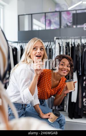 giovani amici multietnici che tengono tazzine di carta e puntano all'abbigliamento in boutique Foto Stock