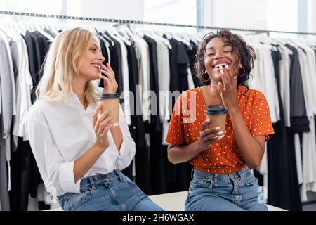 amici multietnici felici che tengono tazze di carta e ridono in boutique Foto Stock