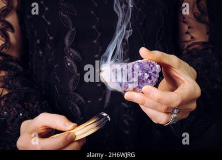 Vista ravvicinata della donna in abito in pizzo nero, pietra miliare in cristallo di ametista da macchiarsi bastone di legno Palo Santo. Rimuovere l'energia negativa. Foto Stock