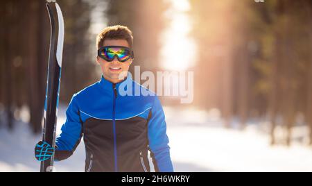 Ritratto felice atleta maschile con sci di fondo in mani e occhiali, allenamento nella foresta innevata. Banner inverno sano. Foto Stock