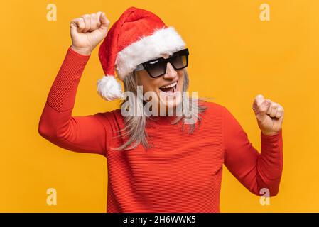 Felice donna di mezza età grigio-capelli in rosso santa cappello e occhiali da sole che ballano, fa movimenti alla musica, sorride positivamente, essendo in alto spirito. Spensierate danze mature della signora isolate su giallo Foto Stock