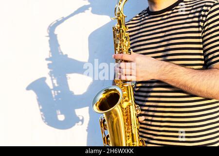 Giovane uomo in un luogo soleggiato all'aperto che gioca sassofono di fronte a un muro. Foto Stock