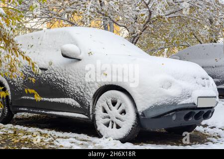 Un'auto grigia coperta con la prima neve sullo sfondo di alberi gialli. Prima nevicata. Problemi per i conducenti in inverno. Cattive condizioni meteorologiche. Foto Stock