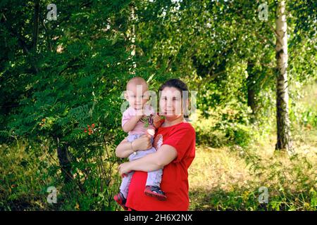 vista di mezza lunghezza di una giovane donna con un bambino nella foresta in piedi sullo sfondo di alberi verdi betula pendula e sorbus aucuparia Foto Stock