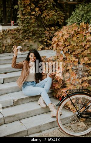 Bella giovane donna con telefono cellulare bere caffè per andare alle scale in bicicletta il giorno d'autunno Foto Stock