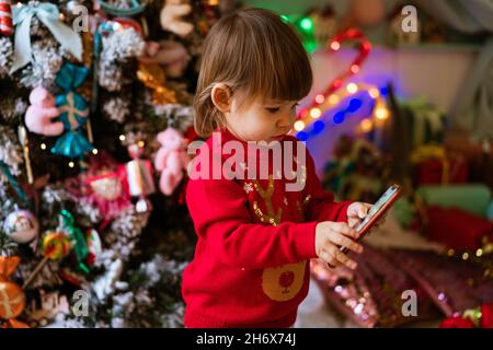 Bambina in maglione rosso sta guardando cartoni animati al telefono accanto all'albero di natale a casa mentre si siede sul pavimento. Bambino in attesa di ricevere regalo per natale e Capodanno. Concetto di tecnologia e bambini Foto Stock