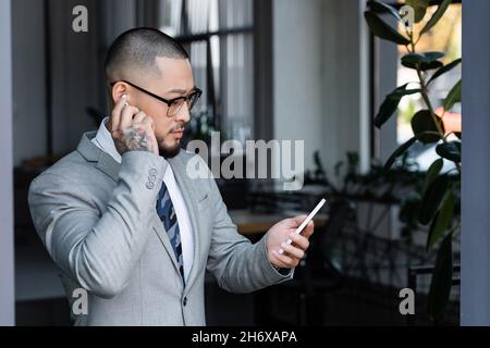 tattoooed uomo d'affari asiatico che inserisce l'auricolare mentre chiama sul cellulare in ufficio Foto Stock
