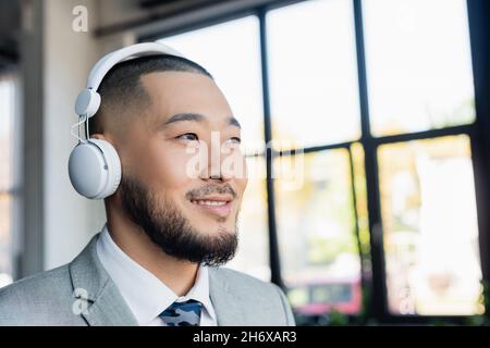 felice uomo d'affari asiatico in cuffie wireless guardando lontano in ufficio Foto Stock
